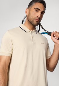 Beige polo tričko s černobílými akcenty na límci, krátkými rukávy a vyšitým logem značky. Model drží tenisovou raketu.