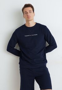 Tummansininen collegepaita, jossa on pyöreä kaula-aukko ja "TOMMY HILFIGER" -logo. Materiaali näyttää pehmeältä ja sileältä. Yhdistettynä vastaaviin sortseihin.