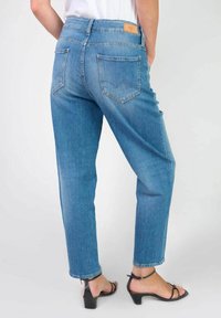Jean en denim bleu clair avec une coupe décontractée, design à cinq poches, et un patch en cuir brun sur la taille, montrant les poches arrière et l'ourlet.