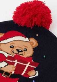 Granatowa dzianinowa czapka z czerwonym pomponem, z motywem niedźwiedzia w stroju Świętego Mikołaja trzymającego prezent. Ozdobiona małymi kolorowymi dodatkami.