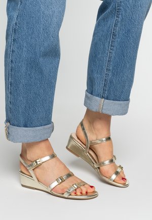 Goldfarbene Keilsandalen aus Metall mit mehreren dünnen Riemen, Schnallen und einer strukturierten Sohle. Kombiniert mit hochgekrempelten blauen Jeans.
