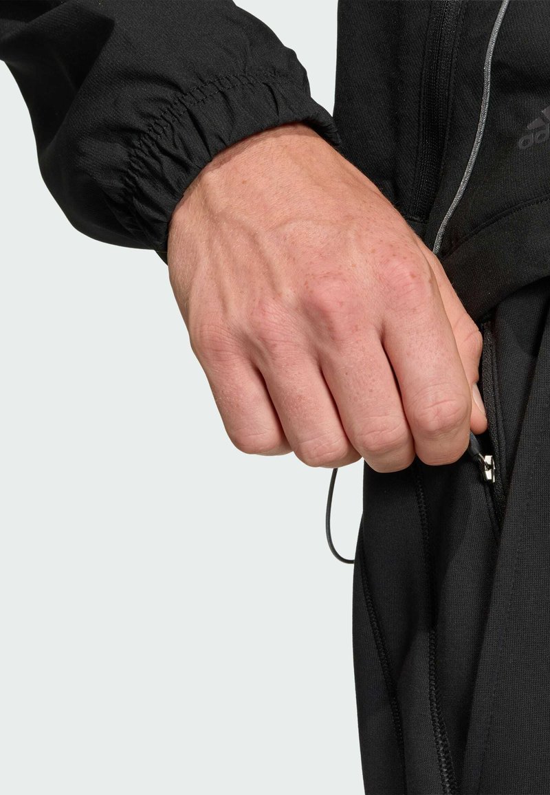 Mano che tiene la linguetta della cerniera nella tasca di una giacca nera Adidas, con polsino elastico e dettagli riflettenti.