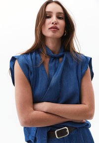 Denimová halenka v tmavě modré barvě s krátkým rukávem, s uzlovým výstřihem a doplněná hnědým opaskem, s jemnou texturou.