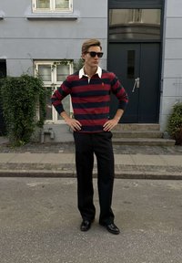 Férfi fekete napszemüvegben, piros és tengerészkék csíkos rögbiingben, fekete nadrágban és mokaszinban, kezét a csípőjén tartva áll az utcán.