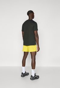 Black short-sleeve t-shirt, yellow shorts, and black sneakers. White socks with logo. Smooth fabric, relaxed fit, side slits on shorts.