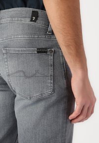 Grå denimjeans med et klassisk fem-lomme-design, en svart logotag på livet, og subtile sømdetaljer på baklommen.