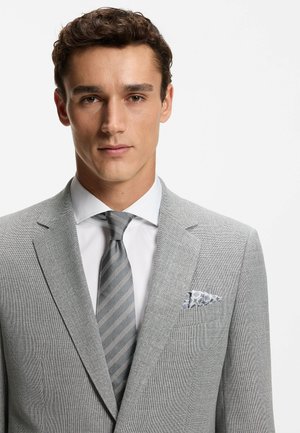 Giovane uomo che indossa una giacca di lino grigio chiaro con trama, camicia bianca, cravatta grigia a righe e fazzoletto da taschino decorato su sfondo bianco.