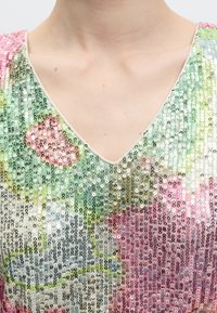 Κοντινό πλάνο ενός ρούχου με V-λαιμόκοψη καλυμμένο με πολύχρωμες παγιέτες που δημιουργούν ένα ανθικό σχέδιο σε ανοιχτόχρωμο δέρμα.