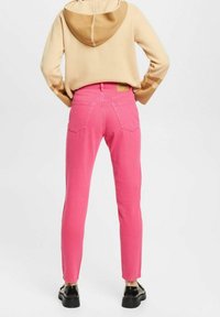 Vaqueros de mezclilla rosa con un corte slim, diseño de cinco bolsillos y un parche de logo en la cinturilla trasera. Llevados con una sudadera beige.
