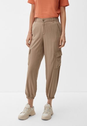 Beige cargobroek met elastische boorden, voorzien van zijzakken en een knoopsluiting, gecombineerd met een roestkleurig t-shirt en beige sneakers.