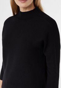 Schwarzer Strickpullover mit hohem Kragen, gerippten Akzenten an den Schultern und einer figurbetonten Silhouette mit Dreiviertelärmel.