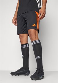 Fekete sportnadrág narancssárga oldalsávokkal, fekete térdig érő zoknikkal, fehér csíkokkal és Adidas logóval, valamint fekete stoplis cipőkkel.