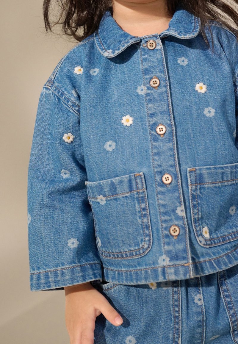 Enfant portant une veste et un pantalon en denim bleu avec de petits motifs de fleurs blanches et des boutons marron, debout avec la main gauche détendue.