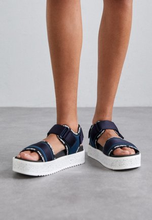 Pieds portant des sandales sportives à plateforme bleu marine avec des sangles ajustables et des semelles épaisses blanches tachetées sur un sol gris.