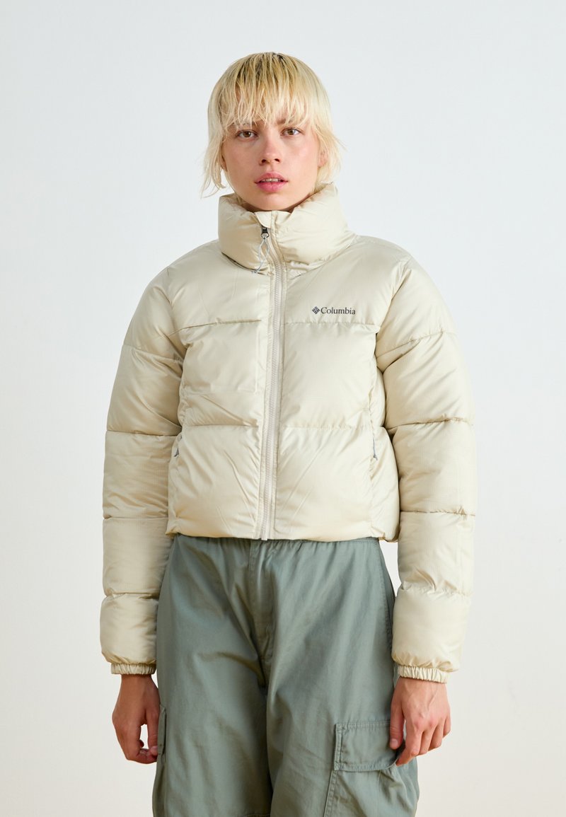 Veste beige rembourrée avec col montant, fermeture éclair sur le devant, et poignets élastiques. Portée sur un pantalon cargo vert olive, avec logo sur la poitrine gauche.