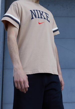 Person, der ein beiges Nike-T-Shirt mit schwarz-weißen Ärmelstreifen und schwarzen Shorts trägt, steht vor einer grauen Wand.