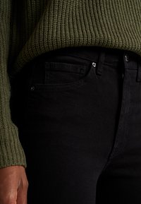 Svarta jeans i smal passform, med fem fickor och silverfärgade detaljer. Den texturerade gröna stickade tröjan täcker delvis midjebandet.