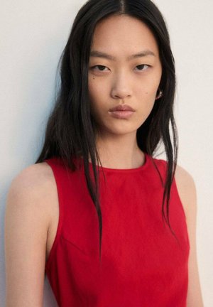 Top rojo sin mangas con escote redondo, textura suave y diseño minimalista. La modelo tiene el cabello largo, liso y oscuro, y lleva pendientes sutiles.