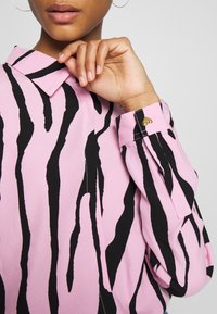 Chemise rose à rayures zébrées noires, dotée d'un col pointu et de poignets boutonnés ornés de boutons en forme de cœur. Tissu à la texture lisse.