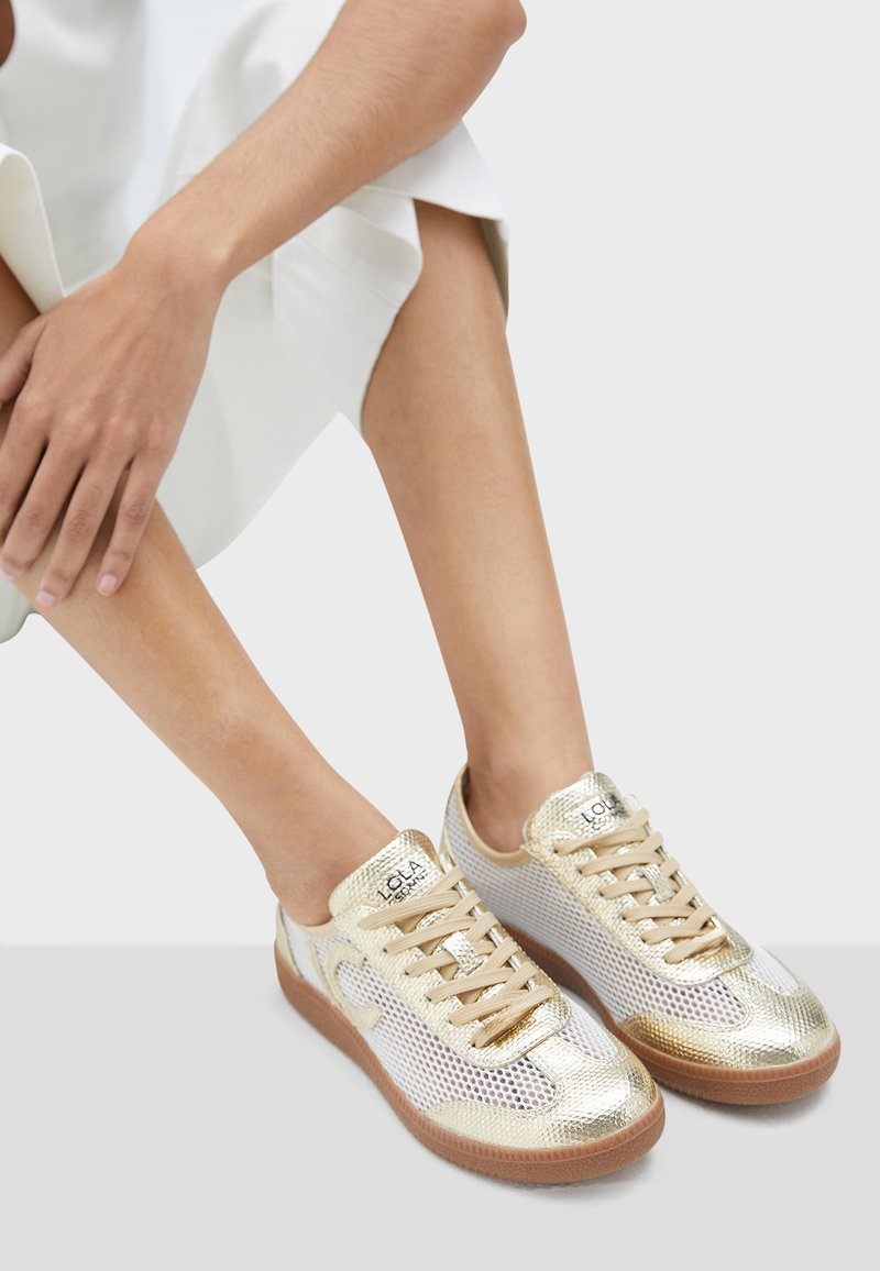Metalické zlaté tenisky s síťovanými detaily, kulatou špičkou a béžovými tkaničkami. Gumová podešev a značka na jazyku.