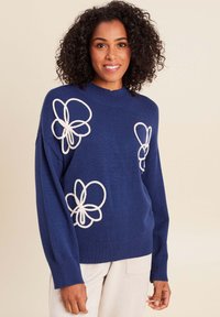 Pull en tricot navy avec des appliqués floraux blancs, des poignets côtelés et un col montant. Texture douce et coupe décontractée.