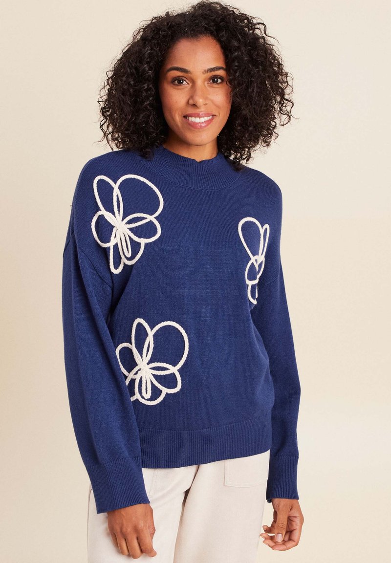 Pull en tricot navy avec des appliqués floraux blancs, des poignets côtelés et un col montant. Texture douce et coupe décontractée.