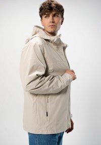 Vedenkestävä vaaleanbeige takki hupulla, jossa on etuvetoketju, sivutaskut ja joustavat hihansuut; sileä pinta ja minimalistinen muotoilu.