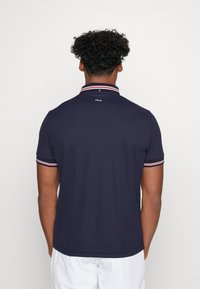 Marineblauwe polo met een klassieke kraag, voorzien van rode en witte strepen, korte mouwen en een subtiele merklogo op de achterkant. Gladde textuur.