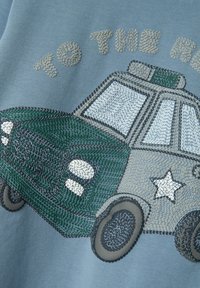 Vaaleansininen kangas sisältää brodeeratun vihreän ja harmaan poliisiautokuvion, jossa on tähti ja yksityiskohtainen etusäleikö. Teksti "KOHTI" auton y above.