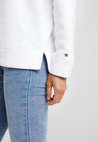 Biała bluza z okrągłym dołem i bocznymi rozporkami, z małym haftowanym logo na mankiecie. Połączona z jasnoniebieskimi dżinsami.