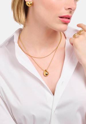 Femme portant des boucles d'oreilles en forme de cœur en or, des colliers en or superposés avec un pendentif en forme de goutte en or et des bagues en or, habillée d'une chemise blanche.