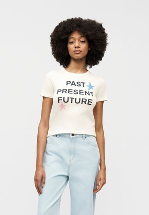 Jeune femme aux cheveux bouclés portant un t-shirt blanc avec le texte "PASSÉ PRÉSENT FUTUR" et des étoiles, assorti à un jean bleu clair.