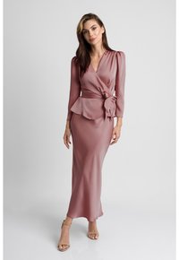 Długa, różowa sukienka z satyny o kroju kopertowym, długimi rękawami, peplum w talii i detalem w formie węzła. Gładka tekstura, sięgająca podłogi sylwetka.