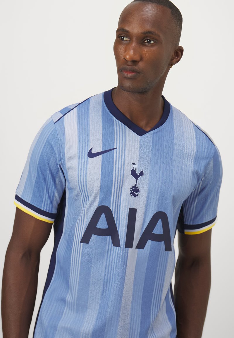 Mężczyzna w jasnoniebieskiej koszulce piłkarskiej Tottenham Hotspur z ciemnoniebieskimi i żółtymi rękawami, spoglądający w bok.