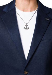 Granatowa marynarka o fakturowym wykończeniu, zapinana na jeden guzik, z metalowym wisiorkiem w kształcie kotwicy na łańcuszku, nałożona na gładką białą koszulę.