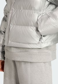 Stříbrná péřová bunda má lesklou, prošívanou texturu s horizontálními švy. Pod ní se nachází měkká, heatherová šedá mikina s třpytivým lemem.