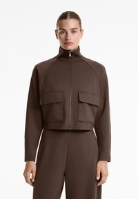Vrouw in een bruine lange mouwen zip-up jack met hoge kraag en grote voorklepzakken, gecombineerd met bijpassende broek.