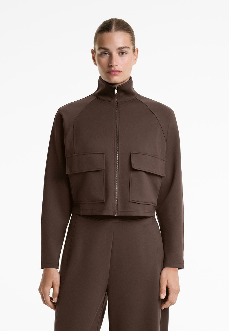 Vrouw in een bruine lange mouwen zip-up jack met hoge kraag en grote voorklepzakken, gecombineerd met bijpassende broek.