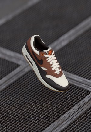 Brown, white, and black Nike sneaker with orange logo, white laces, and gum sole floating above metal grate flooring.