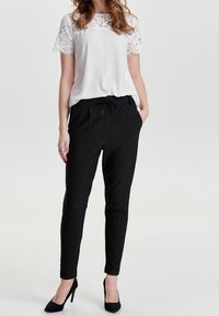 Witte top met kant mouwen, gecombineerd met zwart gestreepte tapered broek. Model draagt zwarte hoge hakken. Lichte achtergrond.