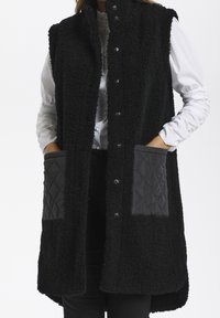 Gilet in pile nero con colletto alto, chiusura a bottoni automatici e due tasche frontali a diamante trapuntate. Texture morbida con un design lungo e aperto.