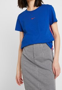 Sininen puuvillapaita, jossa on punainen Nike-logo rinnassa, yhdistettynä musta-valkoiseen houndstooth-kuvioiseen kynähameeseen.