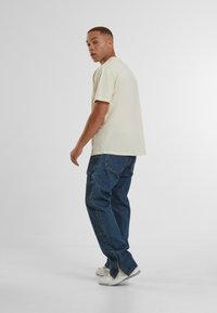 Beige short-sleeve t-shirt and blue baggy jeans. Model stands with back turned, wearing light sneakers. Plain background.