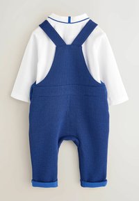 Blaue Latzhose mit einem strukturierten Muster, Trägern und einem weißen Langarmshirt mit einem blauen Kragen. Mit umgeschlagenen Bündchen an den Knöcheln.