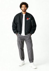 Veste bomber noire avec un motif de cœur rouge et une épingle de sûreté, associée à un pantalon cargo gris et des baskets blanches. La veste est dotée d'une fermeture éclair.