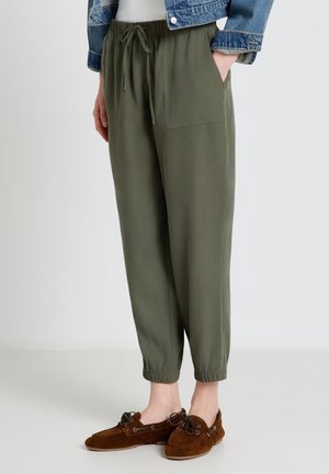 Donna che indossa pantaloni verde oliva con coulisse e polsini elasticizzati, mocassini marroni e una giacca di jeans corta. Mani parzialmente nelle tasche.