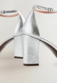 Sandales à talons hauts argentées avec une finition métallique brillante, présentant un design arrière pointu, un talon épais et une sangle de cheville réglable.