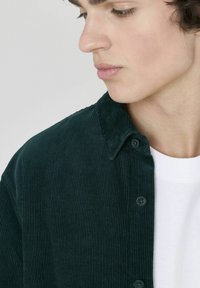 Chemise en velours côtelé vert foncé avec des côtes verticales, col boutonné et deux boutons visibles à l'avant, portée sur un t-shirt blanc.