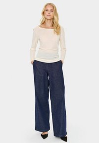Leichtes, langärmliges Oberteil mit weitem Ausschnitt, kombiniert mit weit geschnittenen, dunkelblauen Denim-Hosen mit Fronttaschen und einer strukturierten Oberfläche.