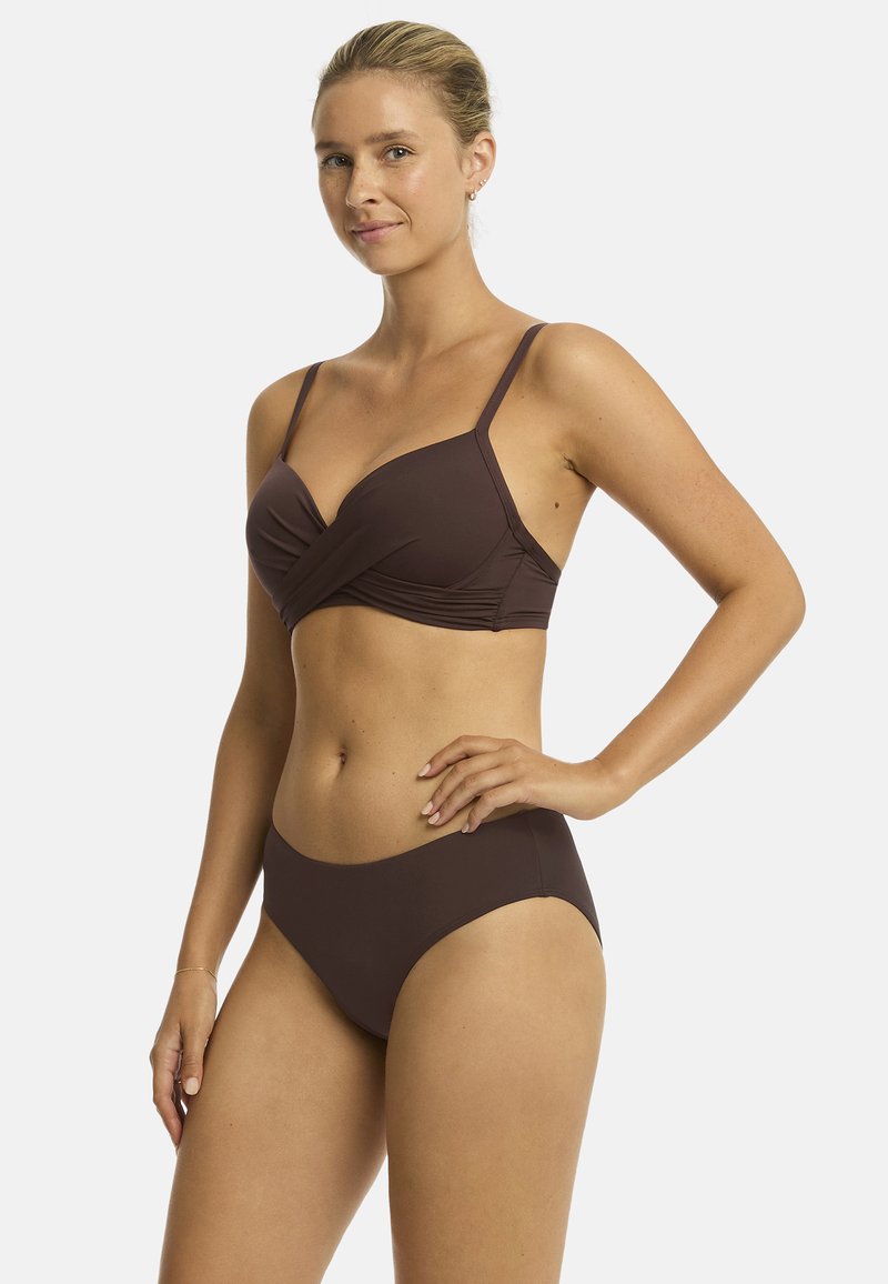 Modelka stoi pewnie w brązowym bikini z wspierającym topem, prezentując zrelaksowany wyraz twarzy na białym tle.